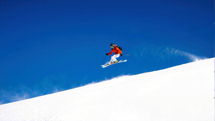 Naklejka na ściany i meble A skier in bright attire is captured mid-jump against a stark blue sky and pristine white snow