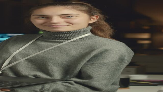 Vertical medium portrait of young Caucasian female programmer posing for camera with laptop while working in dark office alone at night