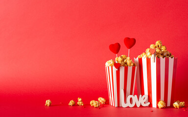 Valentine's Day magic at the cinema. Side view shot captures table set with striped containers holding caramel popcorn, heart ornaments, a love-inspired sign, and sprinkles against a vibrant red wall
