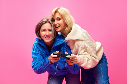 Beautiful young women, lesbian couple playing play station, video games together over pink studio background. Concept of lgbt community, love, Valentine's day, freedom, acceptance, February 14th