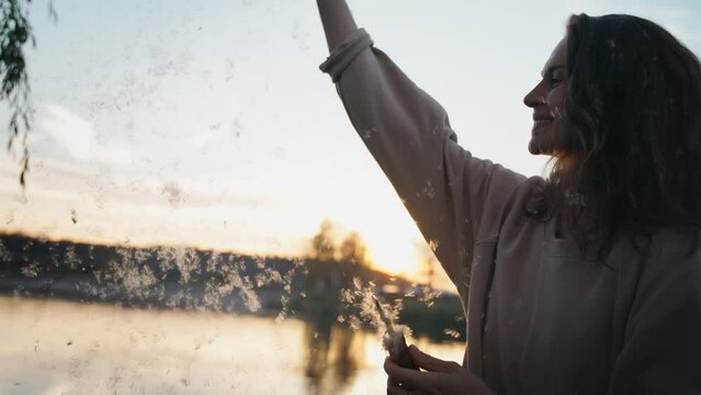 A young cheerful woman having fun playing with down from a bulrush seed head at sunset.