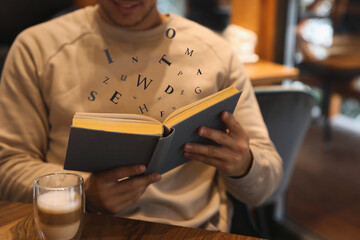 Man with coffee reading book with letters flying over it at wooden table, closeup