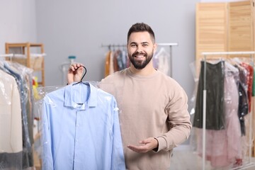 Dry-cleaning service. Happy man holding hanger with shirt in plastic bag indoors