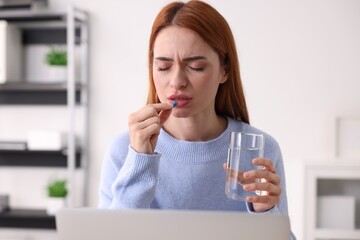 Woman with pill and glass of water suffering from headache at workplace in office