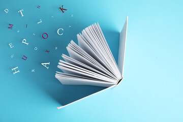 Open book with letters flying out of it on light blue background, top view