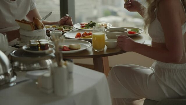 A cinematic shot with unrecognizable faces. A young couple, a man and a woman in white clothes, are sitting having breakfast in a hotel on the top floor of a skyscraper. Healthy meals at the hotel