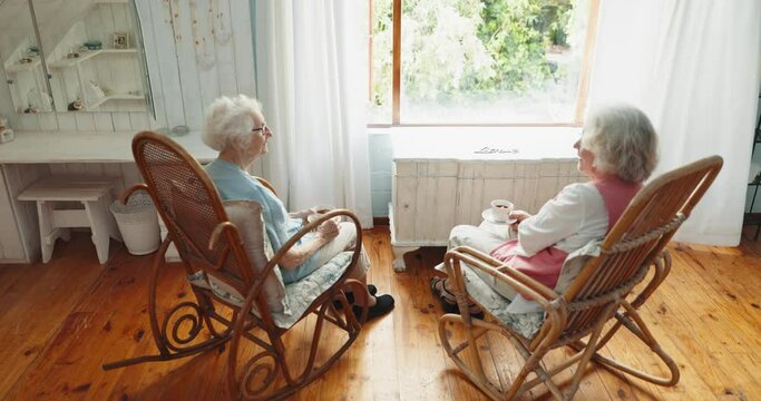 Senior woman, friends and tea cup by window with rocking chair, relax and conversation in retirement. Elderly lady, people and lounge with coffee, coco and chat with memory, thinking and reunion