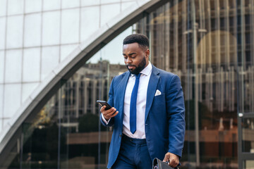 Business black man in suit leaving the office holding his work briefcase and using smartphone