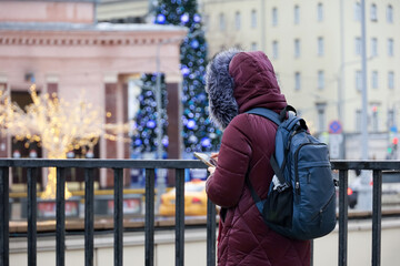 Woman in warm clothes standing with smartphone in hands on winter city street against New Year decorations and illumunation