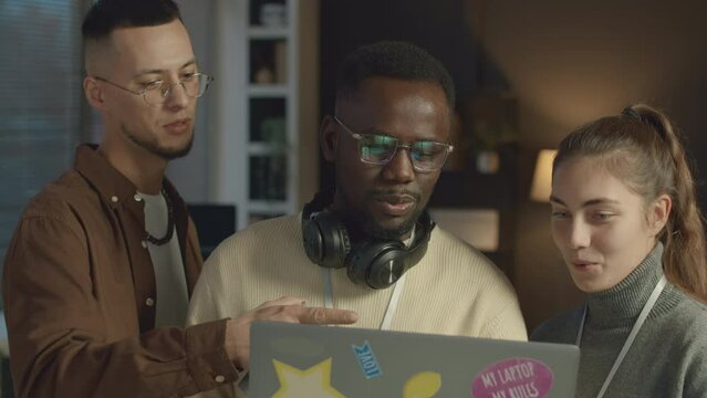 Tilt up shot of two multiethnic men and one woman discussing new software program while standing with laptop in modern programmers office during workday
