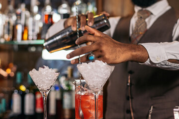 Close-up of the hands of a stylish black bartender preparing a cocktail in a shaker in a...
