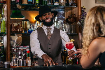 stylish black bartender offering a customer a cocktail decorated with garnish in a traditional...