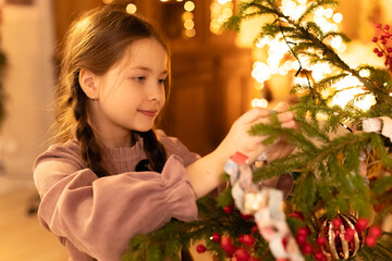 girl with braids decorates Christmas tree with flashing lights by garlands at home during holidays