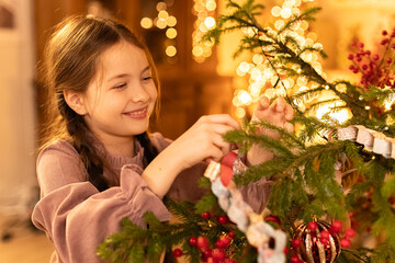 girl with braids decorates Christmas tree with flashing lights by garlands at home during holidays