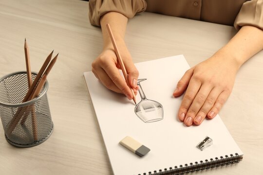 Woman drawing glass of wine with graphite pencil at light wooden table, closeup