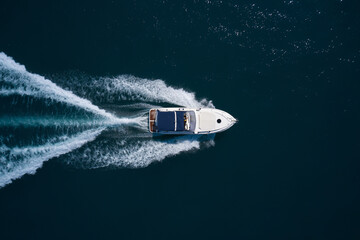 A white boat with a blue awning moves quickly on dark blue water, leaving white traces, top view.