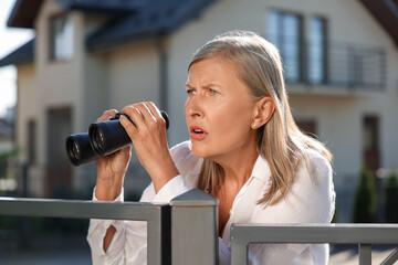 Concept of private life. Curious senior woman with binoculars spying on neighbours over fence...