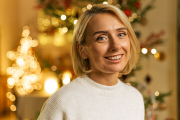 portrait of blond woman smiling near decorated Christmas tree with flashing lights on background