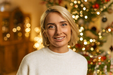 portrait of blond woman smiling near decorated Christmas tree with flashing lights on background