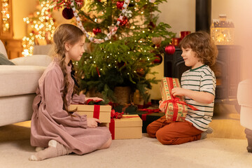 Obraz na płótnie Canvas boy and girl sit near decorated Christmas tree, take presents and shake them to guess what is inside, laugh smile and are excited of foretaste