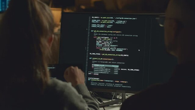 Back view chest up shot of man and woman discussing program code on desktop computer while working in programmers office together at night