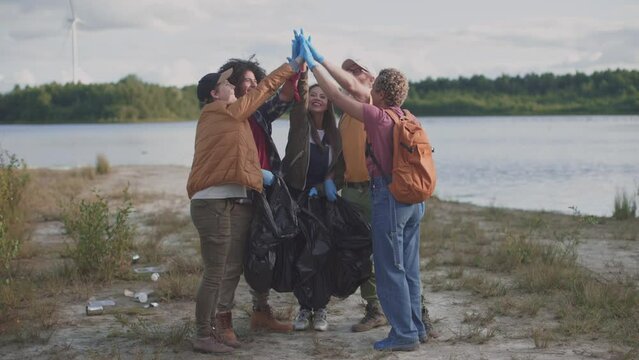 Volunteer team giving high five in nature environment cleaning riverbank from rubbish with plastic bags and gloves