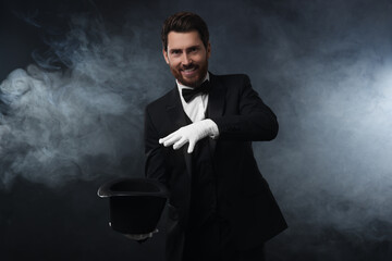 Happy magician showing magic trick with top hat in smoke on dark background