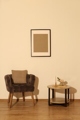 Comfortable armchair, cushion, table and decor elements in room with beige wall. Interior design