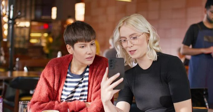 Two Caucasian women resting while sitting in cafe or pizzeria. Young female with blond hair holding phone and showing something to her friend. Happy girls smiling while looking at camera phone.