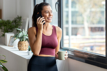 Sporty woman using mobile phone while drinking a cup of coffee after training at home..