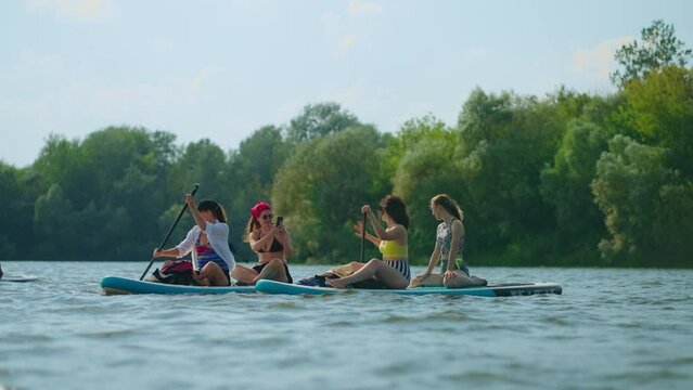 Happy Female Friends Floating On Paddleboard In River And Taking Pictures By Smartphone