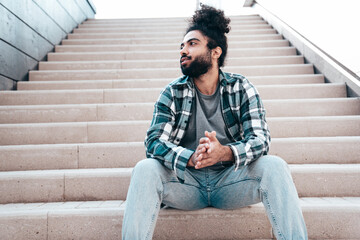 Handsome smiling hipster model. Unshaven Arabian man dressed in summer casual clothes, jeans and shirt. Fashion male with long curly hairstyle posing in the street. Sitting at the stairs - Powered by Adobe