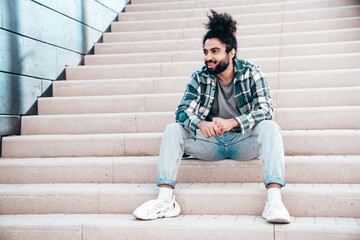 Handsome smiling hipster model. Unshaven Arabian man dressed in summer casual clothes, jeans and shirt. Fashion male with long curly hairstyle posing in the street. Sitting at the stairs