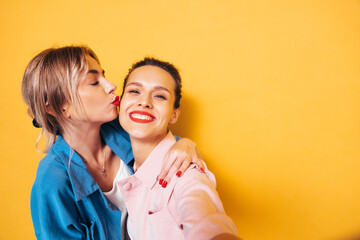 Two young beautiful smiling brunette hipster female in trendy summer clothes. Sexy carefree women posing near yellow wall in studio. Positive models having fun. Cheerful and happy. Take Pov selfie