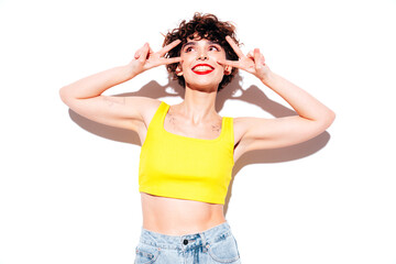 Young beautiful smiling female in trendy summer yellow tank top. Carefree woman posing near white wall in studio with curly hairstyle. Positive model having fun, going crazy. Cheerful and happy