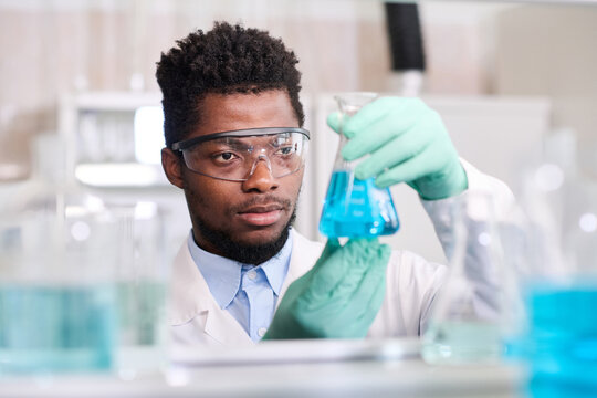 Close up portrait of young African American male laboratory worker in gloves and protective glasses looking at conical flask with blue liquid substance