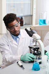 Vertical waist up shot of African American male scientist in safety glasses sitting at desk in...