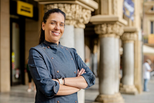 Woman chef wears blue coat, poses with arms crossed and broadly smiles outdoors on the square. Restaurant worker, culinary gourmet, pastry chef.