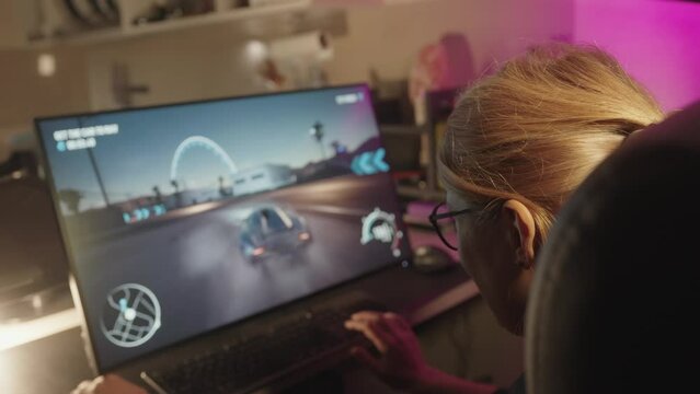 Isolated caucasian gamer girl in glasses sitting in front of a blurred computer screen playing games, focus on players head with screen in bokeh