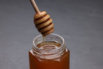 Pouring sweet honey from dipper into jar at grey table, closeup