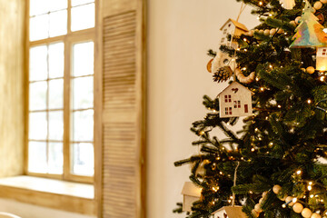 A Christmas tree decorated with wooden ornaments stands near a window with beige shutters