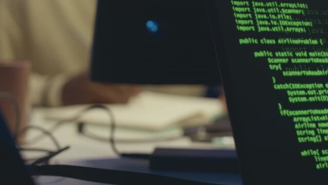 Closeup of green java script code running on black background on laptop and unrecognizable software engineer taking notes in notebook while working in office at night
