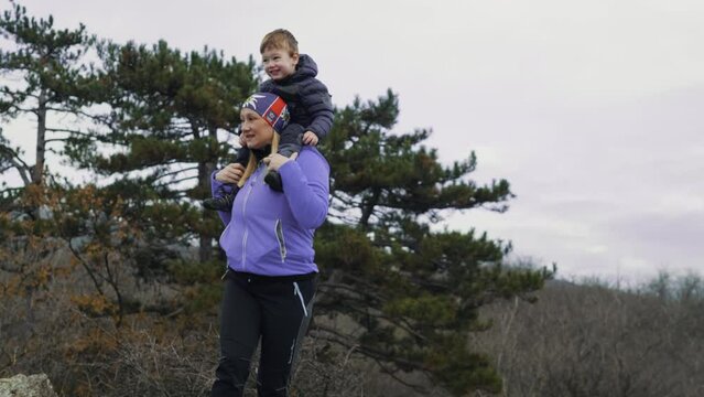 A mother hiker holds her little son on her shoulders and they enjoy looking at the horizon from the top of the mountain