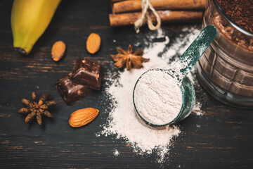 Whey protein powder in measuring spoon on dark wooden board with glass of milkshake smoothie drink, bananas, star anise, almonds, cinnamon stick, chocolate pieces - obrazy, fototapety, plakaty