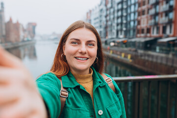 Happy 30s Women taking selfie on urban background. Young beautiful girl say Hi. Gdansk old town and famous Zuraw crane, A beautiful redhead woman is standing near Motlawa river, Traveler