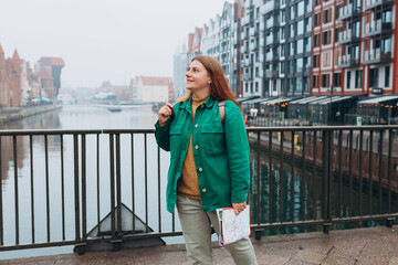 Obraz premium Attractive young female tourist is exploring new city. Redhead girl with backpack holding a paper map on city street in Gdansk. Traveling Europe in autumn. Famous Zuraw crane, Motlawa river