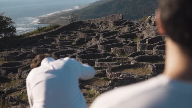 Sunlit summer eve on Santa Tecla, A Guarda. Two young men, one absorbed in the view, the other setting up a camera on a tripod. Ancient Castro, lush hills and Atlantic ocean provide a serene backdrop.