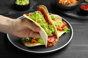 Woman holding taco with guacamole, meat and vegetables at wooden table, closeup
