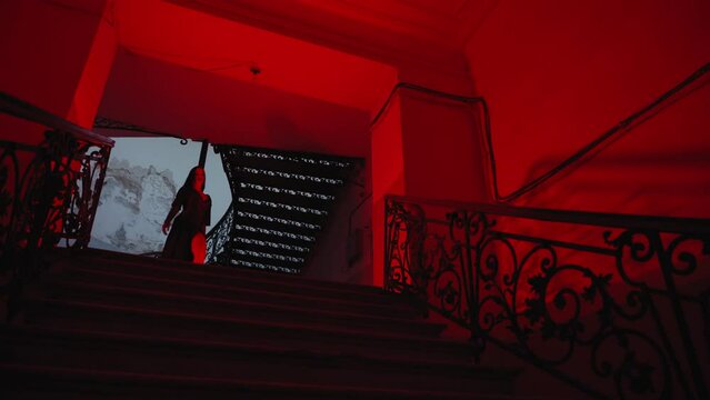 A gothic woman in a black outfit walks down the stairs. The hallway is lit by spooky red lamps, creating a horror movie vibe. It's nighttime indoors.