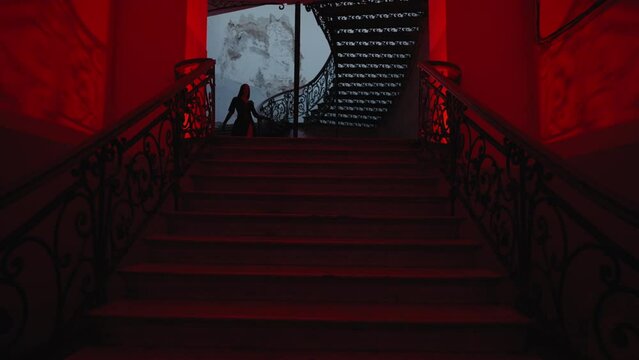 A gothic witch woman in a black outfit walks down the stairs. The hallway is lit by spooky red lamps, creating a horror movie vibe. It's nighttime indoors.